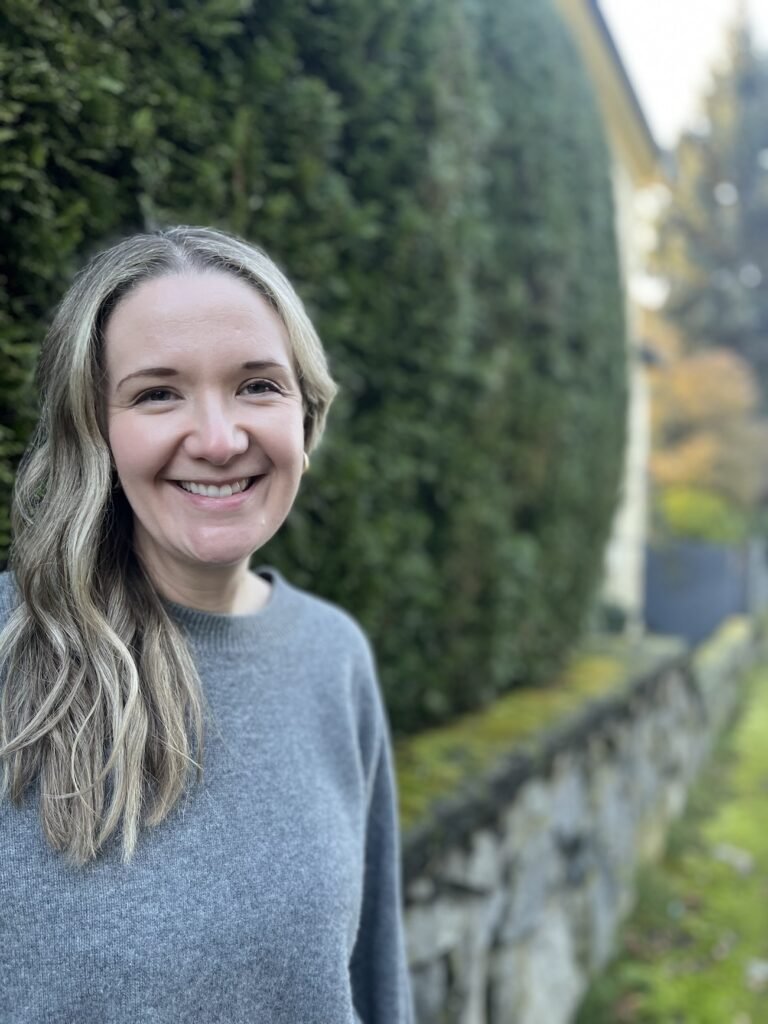 Person smiling outdoors beside a green hedge and stone wall, wearing a grey sweater in soft natural light.