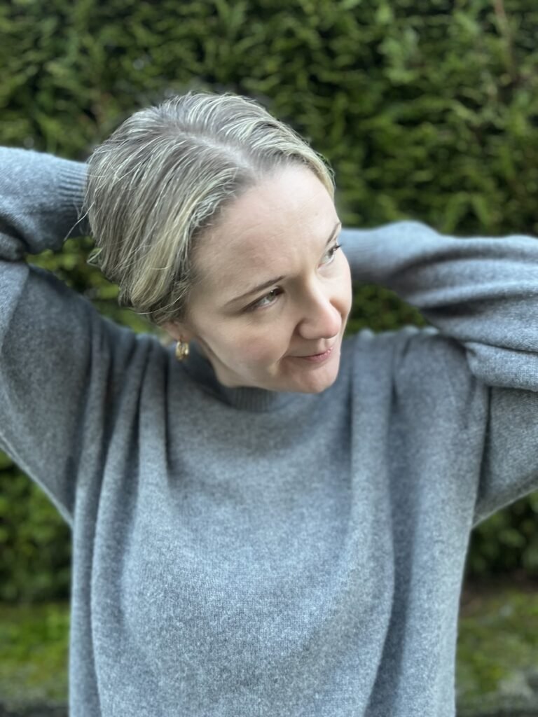 Person standing outdoors in front of greenery, wearing a grey sweater, with arms raised behind their head and a thoughtful expression.